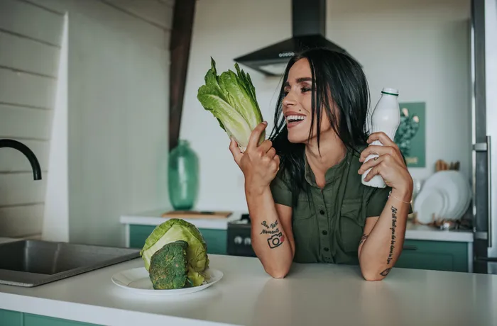 Uśmiechnięta kobieta w kuchni trzymająca sałatę rzymską i butelkę kefiru